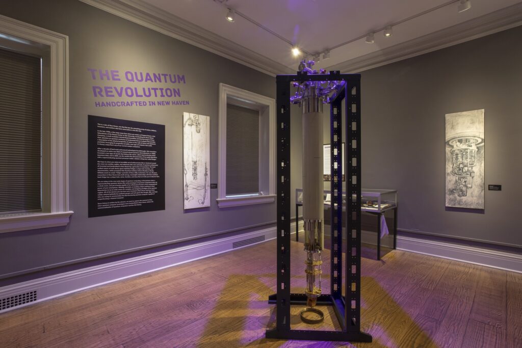 Image of a gallery, with grey walls and a quantum machine/computer on display in the middle. On the left wall is text in purple that says: The Quantum Revolution: Handcrafted in New Haven